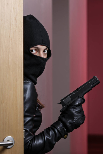 Hausüberfall durch eine Frau in schwarzer Jacke und schwarzer Maske mit schwarzer Waffe. Einbrecher mit Maske. Maskierter Dieb versucht in Wohnungen anderer einzubrechen - Foto, Bild