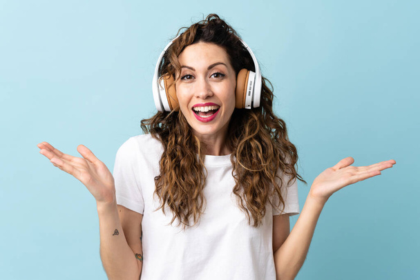 Young caucasian woman isolated on blue background surprised and listening music - Photo, Image
