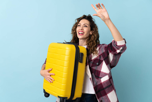Young caucasian woman isolated on blue background in vacation with travel suitcase and saluting - Foto, imagen