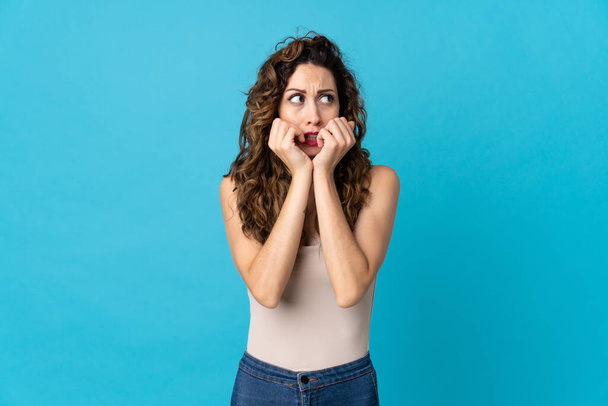 Young caucasian woman isolated on blue background nervous and scared putting hands to mouth - 写真・画像
