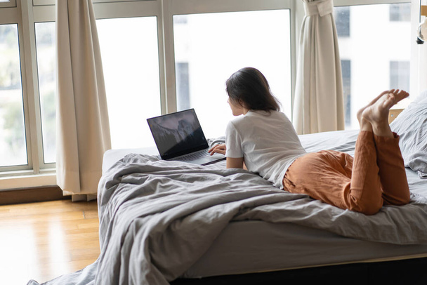 A beautiful young brunette girl is working on the laptop while lying on a bed by a panoramic window. Stylish modern interior. A cozy workplace. Shopping on the Internet - Valokuva, kuva