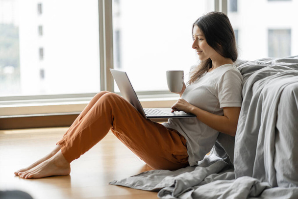Beautiful young brunette girl working on a laptop and drinking coffee, sitting on the floor near the bed by the panoramic window. Stylish modern interior. A cozy workplace. Shopping on the Internet - Foto, imagen