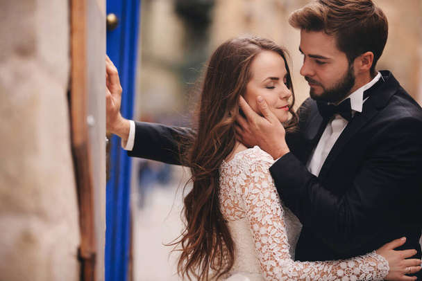 Wedding couple hugging in the old city. Blue vintage doors and cafe in ancient town on background. stylish bride in white long dress and groom in suit and bow tie. wedding day. - Foto, afbeelding
