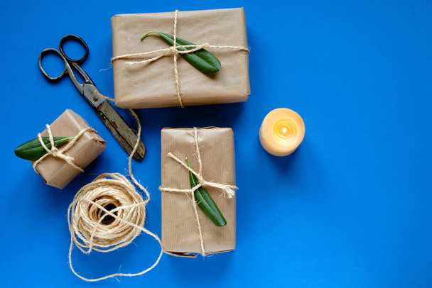 Gift packages in kraft paper, scissors, candle and rope on blue background - Photo, Image