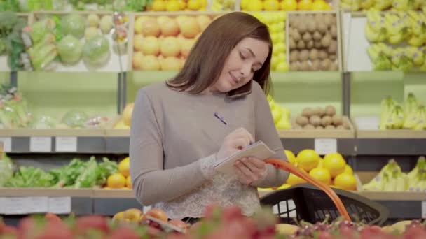 Pretty young woman talking on the phone and taking notes in supermarket. Portrait of charming Caucasian female volunteer clarifying shopping list in grocery. Humanity, lifestyle, volunteering. - Materiał filmowy, wideo