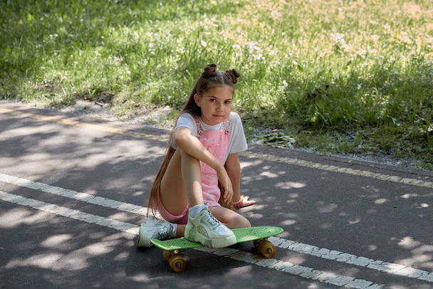 Beautiful girl with long dark straight hair sits on the track with a skateboard. - Фото, зображення