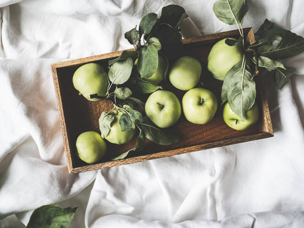 緑のリンゴと白いリネンのテーブルクロスに葉を持つ木製のトレイ。果物の壁紙。トップビュー、フラットレイアウト. - 写真・画像