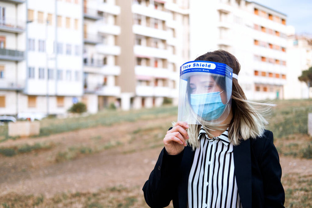Yüzü kalkan izolasyonundaki iş kadını şehir geçmişinde şeffaf bir maske. Sağlık ve sosyal mesafe kavramı. Karantina zamanından sonra.  - Fotoğraf, Görsel