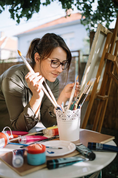 Woman wearing glasses and choosing paint brush from pot - Фото, изображение