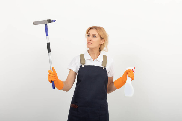 Portrait of cleaning lady or housewife holding a window cleaning brush and a spray. Wearing a uniform apron and latex gloves on light background. European female cleaner ready to work. cleaning agency provides its services - Photo, Image