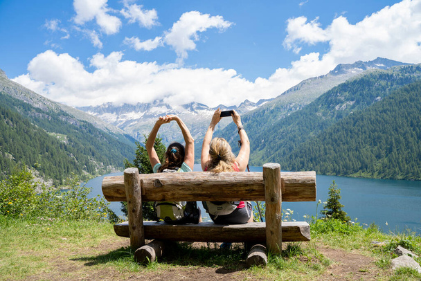 two female friends take a selfie sitting on a wooden bench in front of a mountain lake - Zdjęcie, obraz