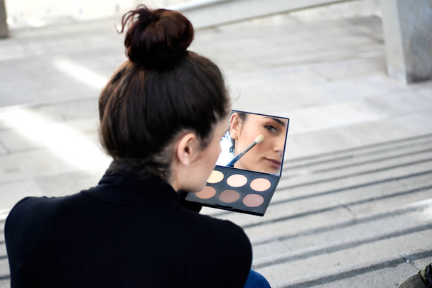 Rear view of a young girl with ponytail sitting on steps outdoors while using a makeup brush - Photo, image