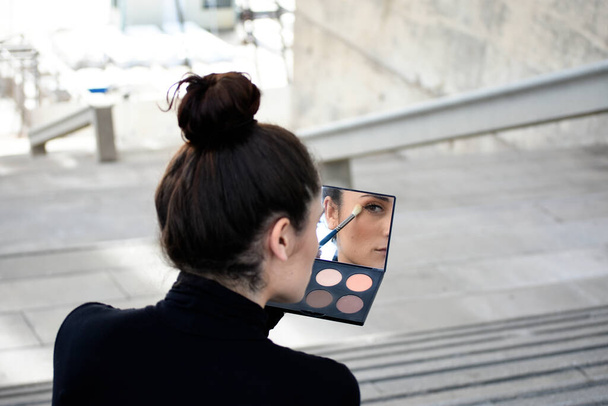 Rear view of a young girl with ponytail sitting on steps outdoors while using a makeup brush - Photo, image