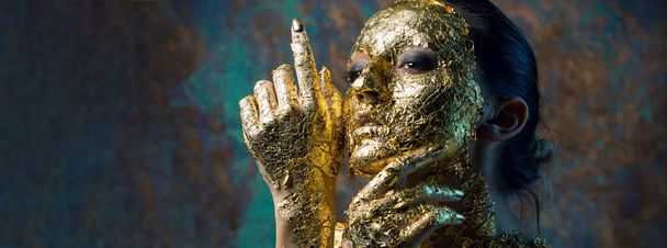 Mädchen mit Maske auf dem Gesicht aus Blattgold. Düsteres Studioporträt einer Brünetten auf abstraktem Hintergrund. - Foto, Bild