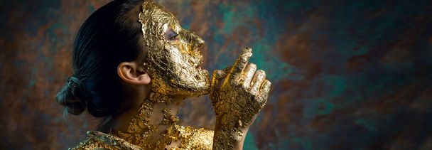 Fille avec un masque sur son visage en feuille d'or. Portrait en studio sombre d'une brune sur un fond abstrait. - Photo, image