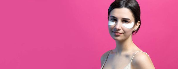 Close up portrait of pretty attractive girl with naked shoulders using patches under eyes. Standing over pink background. - Фото, зображення