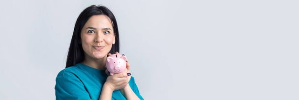 Девушка держит в руках розовую копилку и монету. Концепция богатства и накопления. - Фото, изображение