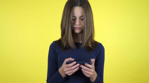 Teenager girl in a blue sweater plays a video game on smartphone on yellow background with copy space. Place for text or product. 4K slowmotion footage - Materiaali, video