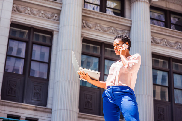 Young African American Woman traveling, working outdoors in New York, standing on street outside vintage office building under sun in summer, working on laptop computer, calling on cell phone - Фото, изображение