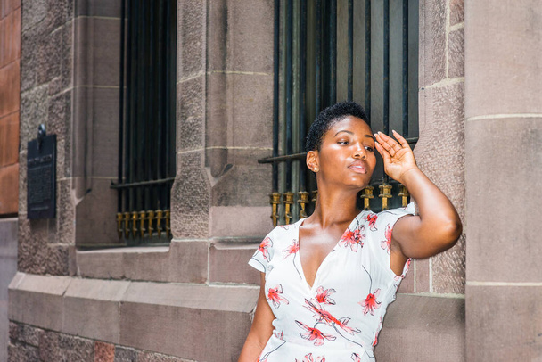 Mujer afroamericana joven relajándose, pensando afuera en la ciudad de Nueva York, vistiendo un vestido blanco con dibujos, levantando el brazo, cerrando la mano a la cabeza, de pie por la pared en la calle, mirando hacia abajo, triste, neumático
 - Foto, Imagen