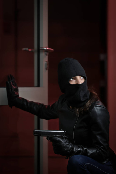 Robo de casa por mujer con chaqueta negra y máscara negra sosteniendo arma negra. Ladrón con máscara. Ladrón en una máscara tratando de entrar en los apartamentos de otras personas
 - Foto, imagen