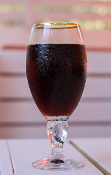 A glass of cold dark beer on a white wooden table in a cafe. The glass is covered with small water droplets. Concept of hot summer thirst quenching, relaxation and fun, drinking beverages  - Photo, image