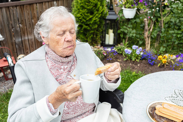 Mulher velha em uma cadeira de rodas bebendo chá no terraço
 - Foto, Imagem