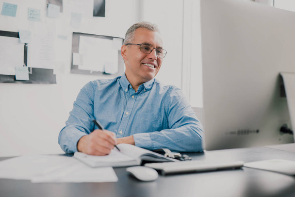 handsome gray-haired smiling man sits in his office while working on business project , checking his laptop while taking notes to his agenda, work routine concept. - Foto, imagen