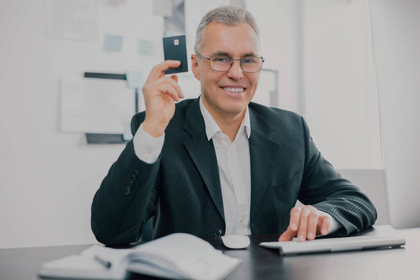 jóképű mosolygós ősz hajú üzletember elegáns öltönyben ül az irodában mutatja bankkártya izgatott, miközben dolgozik az üzleti projekt, pénzcsinálás motiváció koncepció. - Fotó, kép