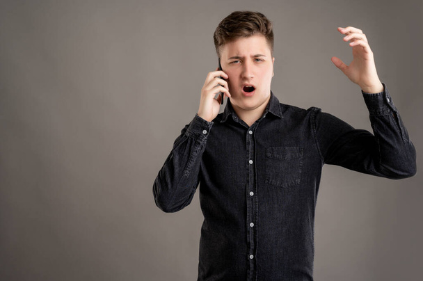 Portrait of serious stylish attractive man dressed with a casual black shirt talking on the phone gesturing angry face isolated on gray background with copy space advertising area - 写真・画像