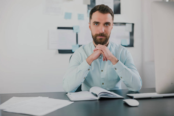 young bearded brunette man looking serious while working on business project in his modern office, holds hands near his face, work routine concept. - 写真・画像
