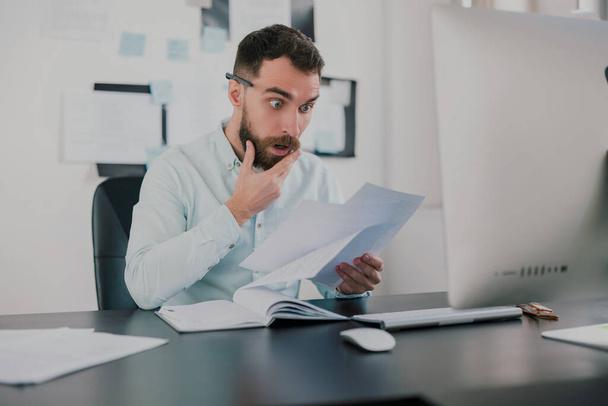 young bearded brunette man looking amazed while working on business project in his modern office, studying documents in his hand, work routine concept. - Foto, Imagem