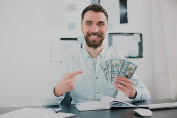 bearded man sitting in modern office holding cash in his hand like card deck looking excited while working on business project , money making motivation concept. - Фото, изображение