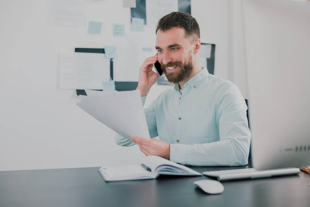 young bearded smiling man working on business project in his modern office, holding documents in his hand, having phone conversation, work routine concept. - Fotoğraf, Görsel