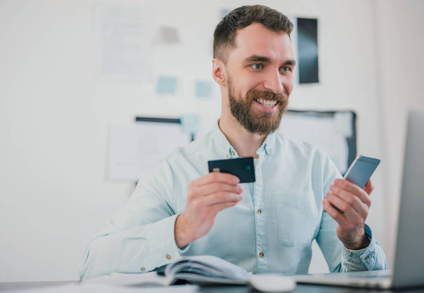 handsome smiling bearded businessman sits in office holding bank card amd smartphone looking excited while working on business project , money making motivation concept. - 写真・画像