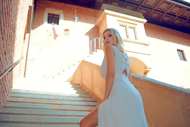 Una mujer elegante en un vestido blanco largo sube las escaleras al castillo. La rubia mira por encima de su hombro a la cámara. modelo en un cuento de hadas
 - Foto, Imagen