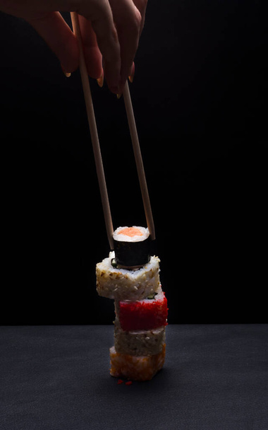 a tower of various Japanese rice rolls and a woman's hand with traditional chopsticks. - Foto, Imagen