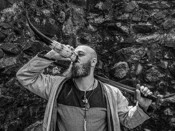 Guerrero vikingo con barba gruesa y grandes bebidas espada del cuerno frente a una pared de piedra, imagen de recreación histórica en blanco y negro
 - Foto, imagen