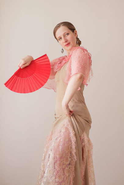 Ritratto di una bella giovane donna in abito beige e rosa su sfondo beige. Sta ballando il flamenco con un fan rosso. Formato verticale - Foto, immagini