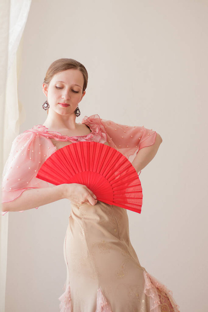 Portrait of a beautiful young woman in a beige and pink dress on a beige background. She is dancing flamenco with a red fan. Vertical format - Foto, Imagem