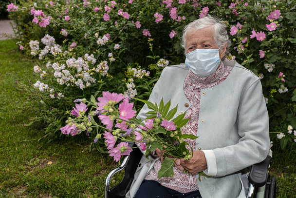 Una anciana en silla de ruedas, en una máscara médica en un paseo por el jardín con flores - Foto, imagen
