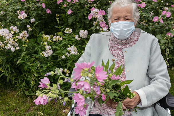 Eine alte Frau im Rollstuhl, mit medizinischer Maske auf einem Spaziergang im Garten mit Blumen - Foto, Bild