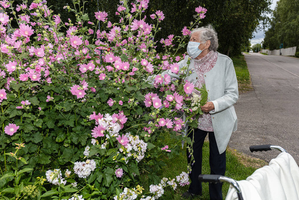 Een oude vrouw in een rolstoel, in een medisch masker op een wandeling in de tuin met bloemen - Foto, afbeelding