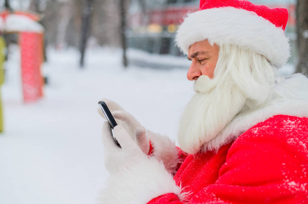 Portrait of a funny funky fat modern Santa Claus use his mobile phone. An elderly man in a suit before Christmas communicates on a smartphone. - Fotografie, Obrázek