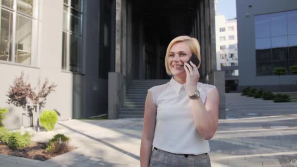 Attractive blonde in a white shirt and a beautiful watch speaks on a mobile phone and smiles outdoors on the background of big office building.Blonde Girl. Successful Woman. - Кадры, видео
