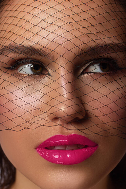 Fashion portrait of a woman's face with evening makeup and a veil on her face. Black mesh and eyeliner, pink full sexy lips and open mouth. White teeth and beauty concept. Clean fresh face skin - Zdjęcie, obraz