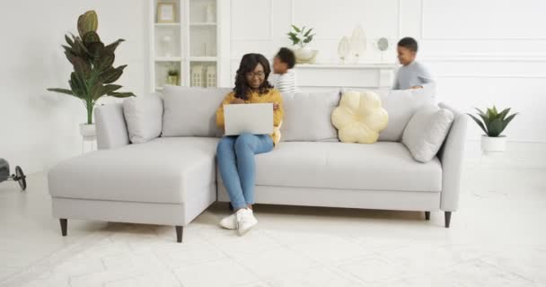 Két boldog afro-amerikai gyerek szaladgál körbe-körbe a nappaliban. Boldog aranyos kisfiú és lány játszik körül kanapé. Mosolygó anya ül a kanapén, dolgozik laptop számítógép otthon - Felvétel, videó