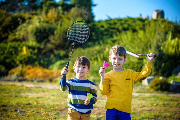 Two active preschool boy playing badminton in outdoor court in summer. Kids play tennis. School sports for children. Racquet and shuttlecock sport for child athlete. Friends happy together warm field. - Photo, Image