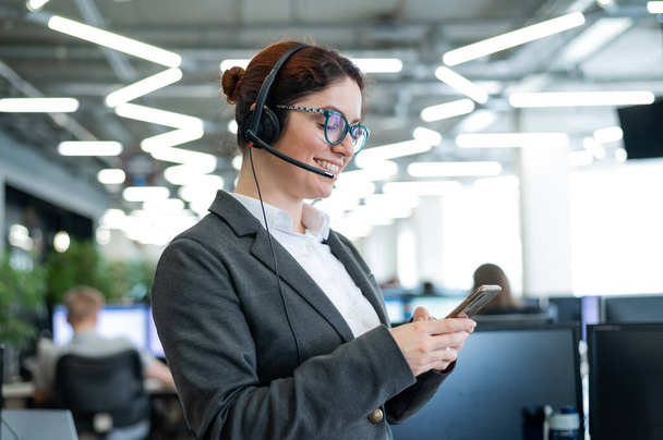 Beautiful caucasian woman in headset is holding a mobile phone while standing in open space office. Friendly female helpdesk operator browsing the screen of a smartphone in the workplace. - Фото, изображение
