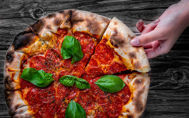 woman Hand takes a slice of Pepperoni Pizza with Mozzarella cheese, salami, Tomatoes, pepper, Spices and Fresh Basil. Italian pizza on wooden table background - Foto, Bild